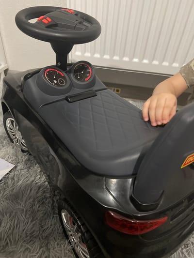 The image shows a close-up of a black ride-on toy car, possibly for a child. The car has a steering wheel with red buttons, two dashboard gauges, and a textured seat. A small child's hand is visible, touching the seat. The car is positioned on a fluffy gray rug. In the background, there appears to be a white radiator or heating unit against a light-colored wall. There is no discernible information to indicate the location is Yekaterinburg, Russia.
