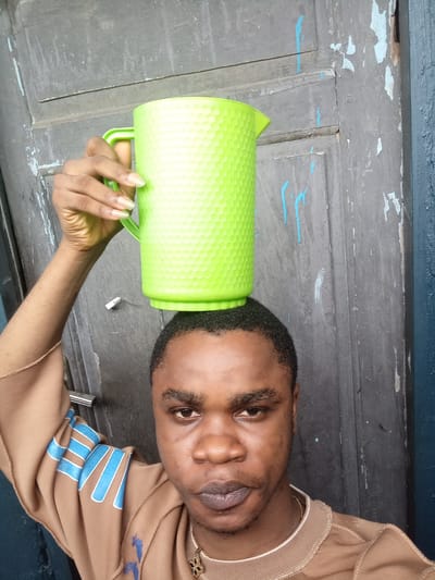A man is holding a green plastic pitcher on his head. He is wearing a brown sweatshirt with blue stripes on the sleeves and a gold chain around his neck. He is standing in front of a gray wooden door with peeling paint. The location is likely Owerri, Nigeria.