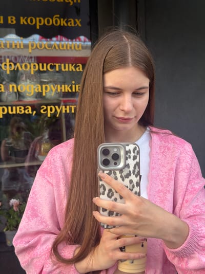 A young woman with long brown hair is taking a selfie with her phone. She is wearing a pink, textured jacket over a white top. The phone she is holding has a leopard-print case. She is also holding a cup in her other hand.
Behind her, through a glass surface, there is text in yellow lettering. The text appears to be in Ukrainian and reads: "В коробках", "атні рослини", "флористика", "подарунків", "рива, ґрунт". This suggests she is likely in a shop selling plants, flowers, and gifts, possibly a florist or gift shop.
The lighting suggests it is daytime. The scene appears to be an indoor setting, possibly a storefront or a room with a window reflecting the outdoors. The woman has a soft smile on her face, giving a pleasant and relaxed impression.