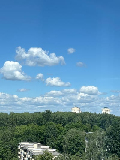 The image depicts a bright, sunny day in Minsk, Belarus, with a clear blue sky dotted with fluffy white clouds. The perspective is from a high vantage point, looking out over a dense, verdant forest.
In the distance, peeking out from above the tree line, are two identical, multi-story buildings. These buildings have a distinctive architectural style, with white facades, numerous windows, and a darker, conical roof. Closer to the foreground, on the right side of the image, a portion of a modern white building with multiple levels and balconies is visible, also nestled amongst the trees.
The thick foliage of the trees dominates the right half of the frame, creating a sense of depth and natural beauty. The sunlight suggests it is daytime, likely late morning or early afternoon, given the brightness and the clarity of the sky. The presence of the buildings interspersed with the natural landscape hints at an urban or suburban environment within Minsk, where residential areas might be integrated with green spaces. There is no text visible in the image. The overall mood conveyed is peaceful and serene, highlighting a pleasant day outdoors.