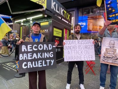 Captured in New York City, United States, the photograph features a group of three individuals engaged in a protest. They are positioned in front of a "KLIKTIX" ticket booth, amidst the bustling Times Square environment. The central figure holds a sign proclaiming, "TODAY, RUSSIA LAUNCHED ITS LARGEST ATTACK ON UKRAINE EVER." To his left, an elderly man stands holding a sign that reads "RECORD-BREAKING CRUELTY" and to his right, a man holds a sign showing a picture of a man, likely Vladimir Putin, with the caption "A WAR CRIMINAL." The Ukrainian flag is seen in the scene, reinforcing the thematic focus of the protest, likely concerning the conflict in Ukraine. Additional individuals and structures visible in the background contribute to the sense of the location being a crowded public space.