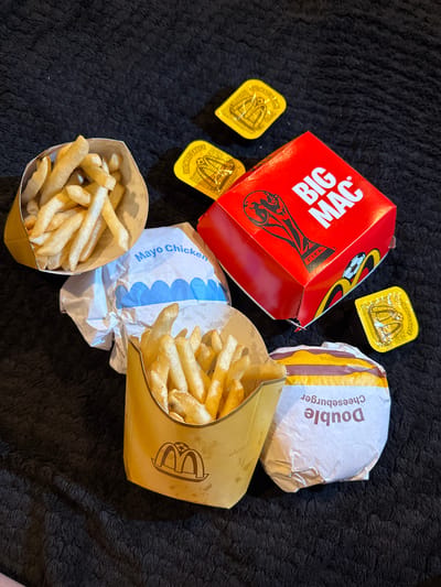 The image displays a spread of fast food items from McDonald's, arranged on a black, textured surface, likely a blanket or rug. The items include two servings of french fries in their signature brown paper containers, one wrapped "Mayo Chicken" burger, one wrapped "Double Cheeseburger", and a red "Big Mac" box. There are also three small, yellow sauce packets, one of which is clearly labeled "Big Mac Sauce". The "Big Mac" box features a FIFA World Cup trophy graphic. The setting is indoors, with no visible windows or external environment. There are no people or animals present in the image. No specific location within Great Yarmouth, United Kingdom, can be identified from the visual information provided.