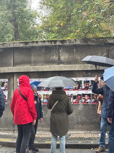 The image depicts an outdoor urban scene in Capital City of Prague, Czechia, under overcast, rainy conditions. Multiple individuals stand in front of a weathered concrete wall, which is largely covered with numerous identical posters.

The environment consists of a textured, light gray concrete wall, approximately 2-3 meters high, topped with a horizontal ledge showing moss growth. Above the wall, dense green foliage of deciduous trees is visible. The ground immediately in front of the wall, partially visible at the bottom, appears to be a paved surface.

At least five individuals are present, most holding open umbrellas.
1.  On the left, a person stands with their back mostly to the viewer, wearing a bright red hooded rain jacket, dark trousers, and dark footwear. A dark shoulder bag strap crosses their back.
2.  To their right, another person faces right, wearing a dark blue jacket, a light blue knit cap, and eyeglasses. They hold a blue umbrella over their head.
3.  In the center, a person stands with their back to the viewer, dressed in a dark olive-green hooded parka/long coat and light blue jeans. A light-colored strap is visible across their back, and they hold a grey umbrella overhead.
4.  On the right, partially visible, a person wears a dark jacket and blue jeans, holding a black umbrella aloft with their right hand.
5.  Further to the right, a person in a brown coat is partially observed.

The concrete wall is systematically covered with white posters, arranged in a grid-like pattern. Each poster features a headshot of a male individual and includes text such as "BRING HIM HOME NOW!" and "KIDNAPPED BY HAMAS," indicating a public awareness campaign for missing persons. The individuals present appear to be observing these posters, utilizing their umbrellas for protection from the visible precipitation. No direct interactions between the individuals are clearly depicted; their focus appears directed towards the wall.
