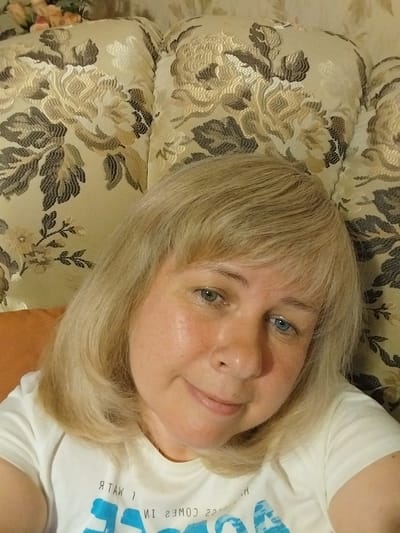 The image is a close-up, forward-facing selfie of a woman with blonde hair and blue eyes. She is wearing a white t-shirt with some text and a blue graphic. The background is a floral patterned couch with a textured fabric. The lighting suggests an indoor setting, possibly a home. The image was taken in Orenburg, Russia.