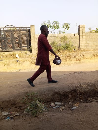 A man walks on a dirt road holding a round, decorated container. He wears a reddish-brown suit with a loose-fitting jacket and pants, and black shoes. The road has a dirt embankment with sparse vegetation and litter. A concrete block wall is visible in the background, along with a decorative metal gate. A few trees dot the landscape. The sky is clear and light, suggesting a sunny day.