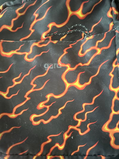 The image shows a close-up view of a black fabric with a flame pattern in shades of red and yellow. The flame pattern is abstract and covers the entire visible surface. A small grey logo, partially obscured by the flames, is visible in the center of the image. The fabric appears to be part of a bag or clothing item, with a visible seam running horizontally across the middle. A thin black cord with white dots is also present, threaded through the fabric above the seam. The image does not provide any information about the location of this item in Gelsenkirchen, Germany, or any context regarding people, actions, or interactions.