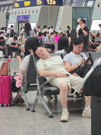This image depicts a group of people waiting in a terminal, likely an airport or train station, in Shangcheng District, China. The scene is brightly lit, suggesting it's daytime. The subjects are seated in rows of gray plastic chairs, some resting their heads on their companions or leaning back, appearing tired or relaxed.
Several individuals are engaged with their mobile phones, while others are looking ahead or conversing. A woman in the foreground wears a light blue shirt and is looking down at her phone. Beside her, a man in a light green t-shirt is asleep, his head resting on the shoulder of a woman in a blue patterned shirt. A bright pink suitcase sits in the lower right corner, suggesting travel.
Visible text includes signage indicating "12B" and "13B" on a blue and yellow striped board, likely gate or platform numbers. Another sign on the wall displays various characters and schedules in Chinese. The overall atmosphere appears to be one of quiet waiting, with people passing time before their departure. The flooring is a speckled gray, typical of public transit hubs. The presence of luggage and seating arrangements strongly indicates a travel-related setting.