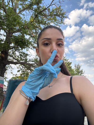 A young adult female is depicted in a close-up selfie, taken outdoors in Kostievo, Bulgaria. She has dark hair, which appears pulled back, and is looking directly at the camera with a neutral expression and slightly pursed lips. Her left hand, covered by a light blue disposable glove, is raised to her face, with the index and middle fingers extended to form a 'V' shape, touching her nose and upper lip. She wears a black spaghetti-strap top, a silver chain necklace with two small heart-shaped charms, and a silver charm bracelet on her left wrist featuring multiple small charms, including an infinity symbol. A small silver stud earring is visible in her right earlobe.
The background consists of a clear blue sky with scattered white cumulus clouds on the right side of the frame. On the left and extending across the top, there is a large, mature deciduous tree with dense green foliage and visible branches. The thick trunk of this tree, located on the far left, has bands of white, teal, and red fabric wrapped around its base. A portion of a green and black patterned folding chair is visible in the lower left, indicating the subject is seated.