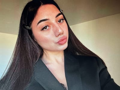 A young woman with long, dark hair and dark brown eyes poses for a selfie. She is wearing a black blazer over a black lace top. Her hair is parted down the middle, and two silver hair clips are visible on the right side of her head. She is wearing light makeup, with emphasis on her lips, which are full and painted with a glossy pink lipstick. Her skin appears fair with some freckles. The background is a plain, light-colored wall with a subtle gradient suggesting lighting. The image is taken indoors, likely in a personal space, given the natural lighting and casual pose. The overall impression is a close-up portrait emphasizing the woman's facial features. There is no information in the image to suggest a location in Chotěšov, Czechia.