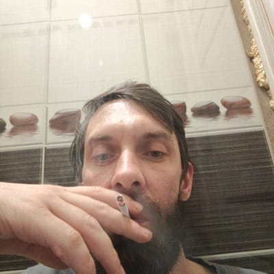 A bearded man is holding a lit cigarette in his hand. He is indoors, against a wall tiled with a pattern of stones. A light fixture is reflected in the tiles. The location is Марковский, Russia.