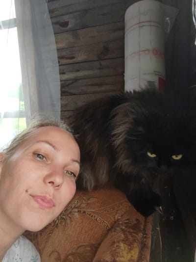 A close-up, selfie-style shot shows a Caucasian woman with blonde hair and green eyes, her face tilted towards the camera. She is positioned to the left of the frame. To her right, a large, fluffy black cat with piercing yellow eyes is visible, its head resting on the woman's shoulder. The background consists of rustic wooden planks, a window with a sheer white curtain on the left, and a roll of what appears to be construction material or plastic sheeting on the right. The woman is wearing a light-colored shirt. The overall setting suggests an indoor environment, potentially a rural dwelling in Чухлэм, Russia.