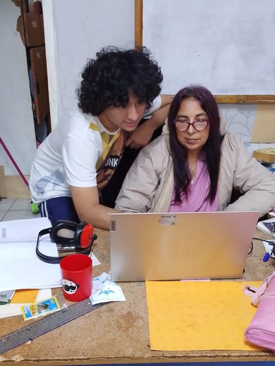 This image captures an indoor scene, likely a home or small office, in Santa Cruz de la Sierra, Bolivia. A young individual with curly dark hair, wearing a white t-shirt with yellow and brown graphic designs, stands behind an older individual. The older individual, with long dark hair and purple streaks, wears glasses with purple frames and a light-colored jacket over a pink garment. Both are looking at a silver laptop positioned on a wooden desk. The desk also displays a red cup with a white cartoon face, a pair of red and black headphones, a notebook, a ruler, and a small package. The background reveals a wall with textured wallpaper and a whiteboard or large piece of paper attached to the wall. The lighting suggests an interior setting, possibly with natural light from an unseen source.