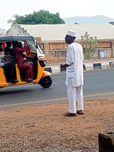 A daylight street scene in Zango-ndaji, Nigeria, depicts a paved road, a gravel shoulder, and background structures under a bright, diffused sky. A Black male, wearing a white cap, dark-rimmed glasses, a white textured traditional tunic with dark blue trim on the sleeves and pockets, and brown sandals, stands on the gravel shoulder in the foreground, facing left. His left arm is slightly bent at the elbow.

On the adjacent paved road, a yellow three-wheeled motorized rickshaw, occupied by at least three individuals, travels from left to right. The driver is visible wearing a maroon-colored long-sleeved garment, steering the vehicle. A passenger in a dark blue shirt and trousers is seated in the front passenger area.

The road features a dark grey asphalt surface, bordered on the right by a white solid line and a black and white striped concrete curb. Beyond the curb, a light brown concrete wall topped with a dark brown metal fence runs parallel to the road. Behind this barrier, a single-story building with a light-colored corrugated metal roof is visible. Green trees and dense foliage, including a palm tree on the left and various bushes, are present throughout the background, with distant hills visible on the horizon. A blue sign with white script is affixed to the metal fence on the right side of the frame. The gravel shoulder consists of small stones and dirt, with a light-colored, elongated object lying on the ground near the standing man's feet. A concrete block is partially visible in the lower right corner of the image.
