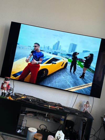 A television screen, tilted slightly, displays a video featuring three individuals and a yellow sports car on an elevated concrete surface with a modern cityscape in the background under a blue sky. The central figure is a male with dark hair, sunglasses, a blue and black patterned shirt, red pants, and tattooed arms, leaning against a bright yellow Lamborghini while giving a thumbs-up gesture. To his right, two females in black outfits are posing; one wears long pants, the other a short skirt and high-heeled boots.
Below the television is a dark entertainment console. On its top surface, from left to right, are a traditional wooden nutcracker figure in red, white, and blue; a small black circular object; several sword-like items, one appearing to be a katana, partially covered by dark fabric; and a small, winged figurine. The console's open shelves contain various items: the upper shelf displays two dark-colored handguns or replicas and several metallic objects; the lower shelf holds an empty clear glass jar, multiple candles in containers, a white decorative sign reading "Always & Forever," a black beanie, and the top section of a black gaming console. Black electrical cables are visible behind and below the television.
The location depicted within the television video, characterized by modern high-rise buildings, is not identifiable as Dresden, Germany; the cityscape is inconsistent with Dresden's typical architecture. The immediate environment where the photograph was taken offers no discernible details to confirm a location in Dresden, Germany.