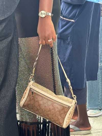 A person with dark skin is wearing a gold wristwatch and a ring on the left hand, holding a Coach handbag. The person is sitting on a seat with a black and silver sparkly covering. Another person is wearing blue jeans and a white and green shoe. A bare foot is visible with a flip-flop sandal.