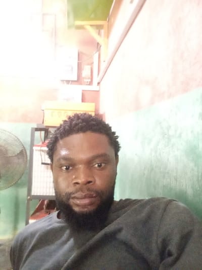 An adult male with dark skin, short curly hair, and a full beard is depicted in a frontal view, looking directly into the camera. He appears to be perspiring, evidenced by a sheen on his forehead and nose. He is wearing a dark, long-sleeved shirt. The scene is an indoor environment, located in Magboro, Nigeria.
The background features a wall partitioned horizontally into two distinct painted sections: an upper reddish-brown hue and a lower faded green. To the left of the subject, the metallic grille and housing of a standing fan are partially visible. Behind the subject and to his left, a metallic shelving unit is present. On its upper tier, a yellow rectangular electronic device, likely a power inverter, is visible. Below it, on the lower tier, a white-covered rectangular object, possibly a battery unit, is stored. Above the shelving unit, on the reddish-brown wall, several electrical components are mounted, including a white rectangular panel, a smaller white meter-like device, and associated wiring. A light-colored wooden structural element extends downwards from the upper right corner of the frame.