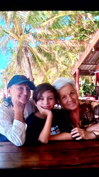 The image shows a medium shot of three people posing for a photo, likely at an outdoor cafe or restaurant. In the foreground, a young person with dark hair and a black t-shirt is positioned between two older women. The person on the left is wearing a blue baseball cap and a white shirt, with their hand resting on their chin. The person on the right has light gray hair and is wearing a patterned shirt, with their arm draped over the young person. They are all smiling and looking towards the camera. The background is blurred, but suggests a tropical setting with palm trees and what appears to be a roof structure. The location is identified as ตำบลเกาะพะงัน, Thailand.
