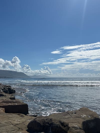 The image displays a sunlit coastal landscape within the Wollongong City Council area, Australia. The foreground features a dark, uneven rocky shoreline composed of angular geological formations, partially wetted by the ocean. The midground is dominated by dark blue ocean water with breaking waves exhibiting white foam. A small, light-colored vessel, containing two human figures whose specific activities are indistinct, is positioned in the water roughly one-third from the left edge of the frame. In the background, a vegetated mountain range extends along the left horizon, with several large white cumulus clouds situated above and partially obscuring the peaks. The sky is predominantly clear blue, transitioning to a lighter tone near the horizon, with additional wispy white clouds in the upper right. Direct sunlight illuminates the scene from the upper right, evidenced by a bright glare.