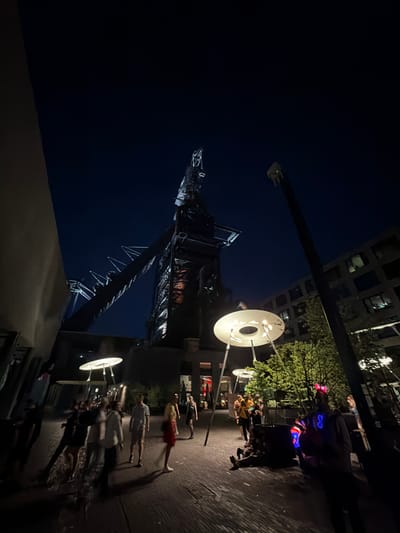 This is a low-angle, nighttime shot of the Belval blast furnaces in Esch-sur-Alzette, Luxembourg. The shot is taken from street level, looking up at the structure. In the right portion of the frame, there is a well-lit area populated with pedestrians. Some are wearing illuminated accessories, like glasses. There are decorative, disc-shaped lights on poles among the people. The building’s infrastructure, including the tower, is a prominent feature. The background sky is dark, indicative of night. Some modern buildings can be seen in the background.