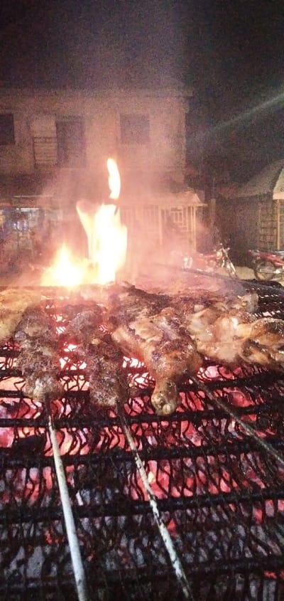 A close-up shot reveals meat grilling over hot coals, the flames rising high, and metal skewers holding the food. In the background, a building is seen with windows. To the side, there's a glimpse of a motorcycle near a small kiosk. The overall scene captures a nighttime barbecue atmosphere with visible fire and grilling taking place.