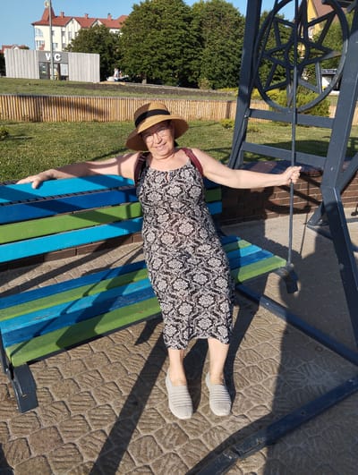 A woman wearing a hat and glasses is sitting on a colorful bench, which is painted in alternating stripes of blue and green. She is wearing a black and white patterned dress and grey slip-on shoes. Her arms are outstretched, and she is smiling at the camera. The bench is located outdoors, possibly in a park or public space. In the background, there are trees, a building with a red roof, and a black metal structure that resembles part of a playground or artistic installation. The ground around the bench is paved with interlocking stones, and shadows from the metal structure fall across it, suggesting it is daytime, likely sunny. The scene appears to be peaceful and relaxed. A sign with "WC" is visible on the building in the background.