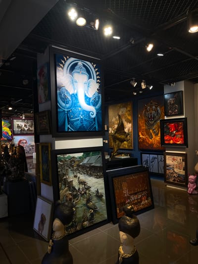 The image shows an art exhibition, likely in Bangkok, Thailand. The exhibition features a variety of artworks, including paintings and sculptures, displayed on walls and shelves. The scene is dimly lit, with spotlights illuminating the artworks. The overall atmosphere is one of quiet contemplation and appreciation for art.
The paintings depict a range of subjects, from a majestic tiger roaring with flames to a serene depiction of a deity with an elephant head surrounded by snakes. Another painting showcases a bustling river scene with numerous boats filled with people, suggesting a vibrant market or daily life in a historical setting. The style of the paintings varies, with some appearing to be realistic and others more fantastical.
Sculptural elements are also present, with at least two figures prominently displayed. One appears to be a stylized representation of a person, possibly a woman, adorned with a golden necklace. Another is a pink pig sculpture, adding a whimsical touch to the collection. The artworks are predominantly framed, with some larger pieces and smaller pieces arranged in clusters.
The setting appears to be an indoor gallery or exhibition space with polished dark floors that reflect the lighting and artworks. The walls are predominantly dark, creating a dramatic backdrop for the displayed pieces. The presence of a metal mesh structure on the ceiling, possibly part of the lighting system, indicates a functional exhibition design.
The image captures a moment of stillness within the gallery, inviting viewers to engage with the diverse artistic expressions. No specific time of day or weather conditions are discernible from the indoor setting. The location context of Bangkok is implied but not explicitly visible through specific landmarks. The emotions evoked are likely those of awe, curiosity, and possibly a sense of cultural richness, given the subject matter of some of the paintings. No visible text is clearly legible within the image that would offer additional information about the artists or artworks.