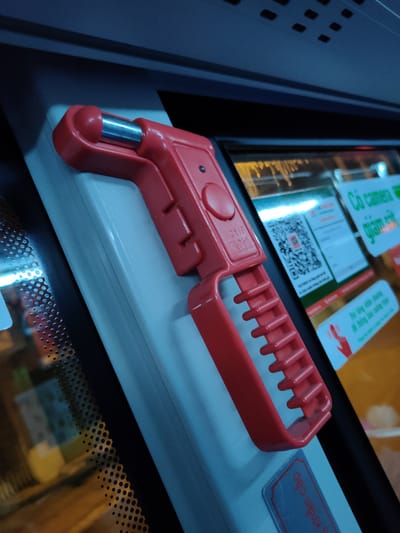 The image shows a red safety hammer with a metal tip, designed for breaking windows in emergencies. It is mounted on a light-colored surface, likely inside a vehicle such as a bus, given the presence of interior elements like a ceiling panel with ventilation grilles and a window reflecting exterior lights. The hammer is positioned at an angle, with its handle featuring a textured grip. To the right of the hammer, a window displays various signs, including a QR code and text in Vietnamese. One sign reads "Có camera" (There is a camera) and "giám sát" (monitoring). Another sign, partially visible, shows instructions related to an emergency, indicated by a hand icon. A blue label with Vietnamese text, "Yêu cầu khẩn cấp" (Emergency request), is visible below the hammer. The lighting suggests it might be nighttime or a dimly lit interior, with some yellow and white lights reflecting on the window, indicating an urban or roadside environment. The overall scene implies a public transport setting equipped with safety features and surveillance.