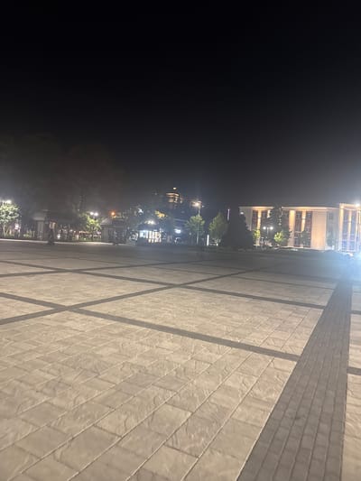 The image depicts a wide, paved plaza at night in Maykop, Russia. The plaza is illuminated by artificial lights, casting shadows from trees and structures onto the light-colored, patterned paving stones. In the background, several buildings are visible. To the left, there are trees and what appear to be small kiosks or structures with lighting. Further back and to the right, a larger, light-colored building with multiple windows and red accents is illuminated. A dimly lit hill with a building on top is visible in the distance behind the trees. Two figures are present on the plaza, one walking in the middle ground and another closer to the background. The overall scene suggests a public space in an urban environment at night.