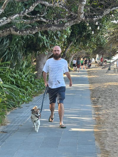 A man with a beard and a white t-shirt walks a small, brown and white dog on a leash along a paved path bordering a sandy beach. He is wearing blue shorts and green flip-flops. Behind him, several other people are walking in the same direction along the path. The path is lined with lush green foliage on the left and trees with hanging lights on the right. The beach to the right of the path is sandy with some lounge chairs visible in the distance. The scene appears to be a tropical location, likely Denpasar, Indonesia, given the vegetation and beach setting. The lighting suggests it is either early morning or late afternoon.
