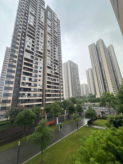 This image depicts a modern residential complex with multiple high-rise buildings. The buildings are characterized by their uniform beige facades and numerous windows, many of which are open or have blinds. Trees and green spaces are visible between the buildings and along a paved walkway. The scene appears to be a residential area, likely in a city.

There are a few people visible in the image. One person is walking on the paved path, holding a blue umbrella, suggesting it might be raining or has recently rained. Another person is visible further down the path. In the distance, there are parked cars and motorcycles.

The sky is overcast and a dull gray, indicating a cloudy or potentially rainy day. The overall lighting suggests it is daytime, but the overcast conditions create a subdued atmosphere. The architecture of the buildings, with their modular design and extensive glazing, points to a contemporary urban setting.

There is some visible text on banners or signs placed along the walkway, appearing to be in Chinese characters. One banner is red and prominent. The location context provided is Chongqing High-tech Zone, China, which aligns with the modern urban development seen in the image.