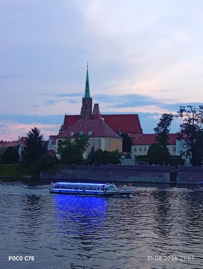 A boat with blue lights is cruising on a river in front of a cityscape. The boat appears to be a passenger vessel with a roof and windows, and it has text on its side that reads "HARBOUR KARDYNALSKA WWW.STATER.COM.PL". In the background, there is a large building complex with red roofs, characteristic of a historic European city. A prominent green spire, part of a church or cathedral, rises above the buildings. The sky is a soft gradient of blue and pink, suggesting it is either early morning or late evening. The river reflects the lights from the boat and the sky, creating a shimmering effect on the water. The overall scene depicts a tranquil view of a European city at dusk, with the river and a boat as the primary focus. The image is located in Wrocław, Poland, specifically along the Odra River with a view of Ostrów Tumski, the historic cathedral island, and the surrounding architecture.