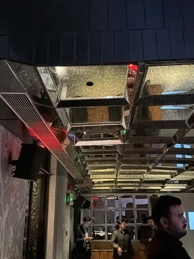 An interior view of a contemporary establishment, likely a bar or restaurant, in New Delhi, India, captured under dim, artificial lighting. The overhead features a striking, multi-faceted mirrored ceiling composed of numerous square and rectangular panels, some displaying a subtle geometric or wavy pattern, interspersed with plain reflective surfaces. Recessed lighting fixtures, including a prominent circular black one, are visible. A long, metallic ventilation grille runs diagonally across the upper left, emitting a distinct red glow. Further into the ceiling, additional red and green indicator lights are discernible. The ceiling's reflective properties create distorted reflections of the room below.
To the left, a light-colored wall with a subtle, intricate white floral or leaf pattern is visible, on which a large, black, rectangular speaker is mounted. A concrete-like pillar stands centrally, separating this wall from a large window-grid structure in the mid-right. A green-illuminated box, possibly an emergency sign, is fixed on a wall section near the pillar.
In the foreground on the right, the profile of a bearded man wearing a dark top is prominent, illuminated by a cool, blue-white light from his right side. Further left, a man in a dark suit, possibly staff, is observed looking down at a mobile phone while standing near a wooden counter. Behind the foreground figures and through the window-grid, several other individuals, including a woman in a dark uniform, are visible, suggesting a moderately populated venue. The overall environment suggests an evening setting within a modern and stylish social space.