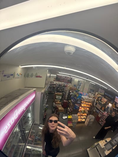 A wide-angle, distorted selfie captures the interior of a Lawson convenience store in Changning District, China, via a convex security mirror mounted on the ceiling. In the immediate foreground, a young adult female with long, wavy brown hair wears dark sunglasses and a dark top. She holds a smartphone, revealing a ring on her right ring finger, as she photographs her reflection. Behind her and to the left, a Haagen-Dazs freezer unit displays a variety of ice cream tubs, with "Haagen-Dazs" visible in reverse on its pink sign. The store's interior features light-colored walls, one of which has a mural depicting stylized buildings, trees, and a red car, along with the reversed "LAWSON" logo. Fluorescent strip lighting illuminates the ceiling, where a white dome-shaped security camera is also visible. Shelves throughout the store are stocked with various products, including packaged foods, drinks, and baked goods in illuminated displays. To the right in the background, a young adult male with dark hair, dressed in dark clothing and white sneakers, stands in an aisle among the merchandise. The floor is a light grey. Chinese characters are visible on product packaging and signage throughout the store.