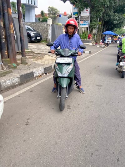 A man in a red helmet sits on a green scooter. The scooter's license plate reads "D 3046 AFC." The man is wearing a blue long-sleeved shirt, blue jeans, and black sandals. The man is facing forward. The scooter is stopped in the middle of a paved road with white lines on either side. The scooter is facing forward. There are several buildings in the background. The image is taken in Bandung, Indonesia, at coordinates (latitude: -6.9, longitude: 107.628).