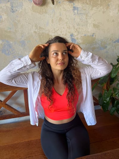 In Phu Thuy Ward, Vietnam, a woman with long, curly brown hair, wearing a vibrant coral-colored sports bra and black leggings, is seated on a wooden bench. She has a white collared shirt draped open over her shoulders, with her hands positioned near her head. The background features a textured wall with a distressed, multi-colored paint finish, exhibiting shades of white, pale blue, and beige. To the right of the woman, a lush green plant with large leaves is visible. The bench itself is constructed of dark, polished wood, featuring a slatted design on its backrest.