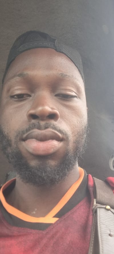 The image shows a close-up, slightly rotated view of a man's face. He has dark skin, a beard, and is wearing a black cap. He is also wearing a red and black shirt with an orange trim, and a brown strap crosses his shoulder. The background is blurry and out of focus, suggesting an interior space. There is no information in the image to indicate that it was taken in Ikeja, Nigeria.