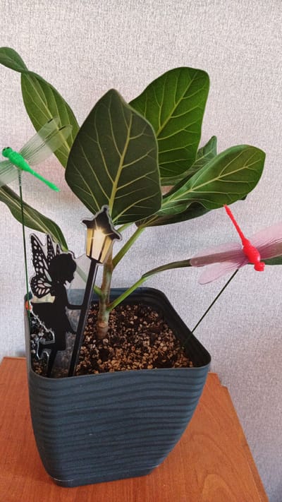 A potted plant with large green leaves is adorned with fairy and dragonfly garden decorations. The plant is in a dark gray pot filled with soil and gravel, placed on a wooden surface. Behind the plant is a textured, light-colored wall. Two dragonfly decorations, one green and one pink, are perched on thin green stems. A black silhouette of a fairy with wings and a small lamppost decoration are also placed in the soil. The scene appears to be indoors, on a table or shelf, with no indication of the specific city of Kryvyi Rih, Ukraine.
