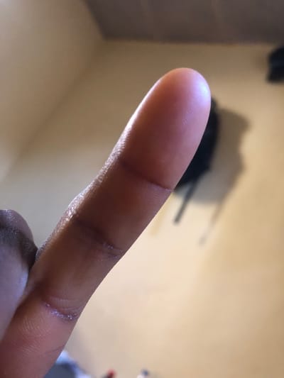 The image displays a close-up, high-angle perspective of a human finger, oriented diagonally from the lower-left to the upper-right of the frame. The finger exhibits dark skin pigmentation, with visible dermal ridges on the fingertip and minor creases at the joint lines. The background is out of focus, indicating an indoor environment with a plain, light brown or beige wall. In the upper-right background, a blurred, dark object resembling a hanging bag or backpack is visible, casting a distinct shadow to its right on the wall. The lighting appears soft, originating from the upper-left, accentuating the finger's contours. The scene lacks specific identifying markers such as architectural features, landmarks, or signage that would allow for the determination of a precise location within the city of Abuja, Federal Capital Territory, Nigeria.