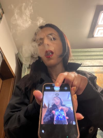 A woman is taking a photo of herself with a smartphone. She is exhaling a cloud of vapor from her mouth, presumably from a vape or e-cigarette. The photo on her phone shows a similar image of her taking a photo, creating a recursive effect. She is wearing a dark-colored jacket. The background appears to be an indoor setting, possibly near a window or light source. The image does not contain enough specific details to accurately identify the location in Konz, Germany.