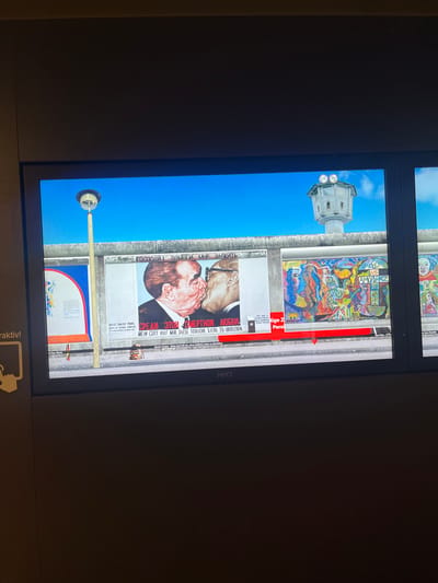 The image displays a digital reproduction of the East Side Gallery in Berlin, Germany. This is a section of the Berlin Wall that has been transformed into an open-air art gallery. The scene shows a vibrant blue sky above a graffiti-covered section of the wall, which features several distinct murals.
Prominently displayed is a well-known mural depicting a fraternal kiss between Leonid Brezhnev and Erich Honecker, leaders of the Soviet Union and East Germany respectively. Below this image, text in Russian and German reads "Gott, hilf mir, diese tödliche Liebe zu überleben," which translates to "God, help me survive this deadly love."
To the right of this mural is a colorful abstract artwork featuring stylized human faces and the word "AMOR-PAZ," meaning "Love-Peace." Further to the right, another mural shows a depiction of a young girl.
In the background, a concrete watchtower, a remnant of the wall's original purpose as a border fortification, stands against the sky. A tall, slender lamppost stands to the left of the murals. The bottom of the display shows the brand name "MMT." The overall impression is that of a historical exhibit, likely in a museum or gallery setting, showcasing a segment of the Berlin Wall's artistic transformation.
