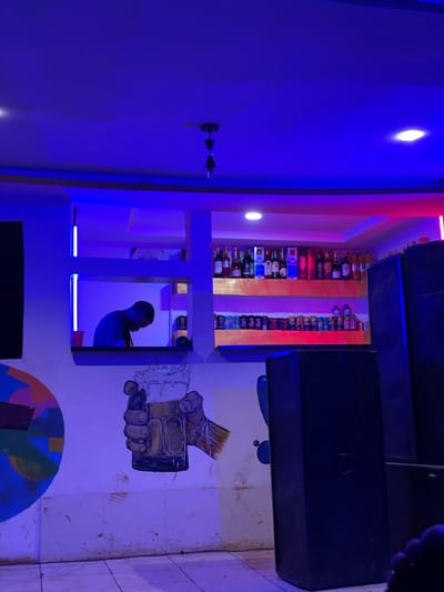 The image captures an indoor scene, likely a bar or a nightclub in Durumi, Nigeria, illuminated by vibrant blue and pink neon lights. The orientation of the image is sideways, suggesting it was taken with a phone held vertically and then rotated.
In the foreground, a large, dark speaker hangs from the ceiling, casting a shadow. To the left, a wall is bathed in intense blue light, with two circular lights on the wall and a two-pronged object mounted on it. Further into the scene, a section of shelving is filled with numerous bottles of alcohol, arranged neatly. Red and blue neon lights add to the ambiance.
A significant portion of the image is dedicated to a large mural on the wall. The mural depicts a hand holding a frothy beer glass. Below this, a colorful abstract design resembling a globe or a circular pattern is visible. In the mid-ground, through an open doorway or alcove, a young man is seen from the side, looking towards the right. He appears to be engaged in some activity within this space, which is also lit by blue neon lights.
The overall impression is one of a lively, modern entertainment venue. The lighting creates a dynamic and somewhat moody atmosphere. The presence of alcohol bottles and a mural suggests a place where people gather to socialize and enjoy themselves. The time of day is indeterminate due to the artificial lighting, but the atmosphere suggests it could be evening or night.