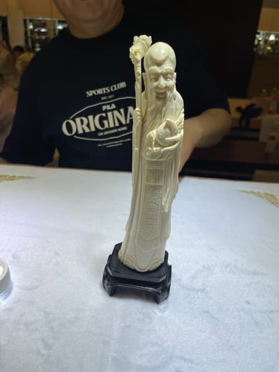 A carved ivory figurine of an elderly man with a long beard and traditional robes stands upright on a black base. The figurine is intricately detailed, with delicate carvings representing facial features, hair, and clothing. The man appears to be holding something in his hands. The figurine is placed on a white, textured surface, possibly a tablecloth or a fine fabric. In the background, a person wearing a black t-shirt with white text is partially visible. The t-shirt has a graphic design and text that reads "SPORTS CLUB," "EST. 1981," and "ORIGINAL FILA UN GRANDE NOME." The scene appears to be indoors, with artificial lighting. The overall mood is calm and focused on the display of the artifact.
