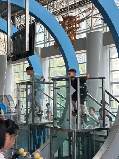 In the city of Jiangbeicheng Street, China, a science museum exhibit is depicted. Two young boys are interacting with a physics demonstration. The boy on the left, wearing a striped t-shirt and denim shorts, is ascending a platform with a metal railing. The boy on the right, in a black t-shirt and shorts, is also on a platform, holding onto the railing. Other children and adults are visible in the background, some observing the exhibit. The exhibit features transparent walls and metal structures, with a large blue curved element overhead and a wooden mechanical model suspended above. Large windows in the background reveal a cityscape with buildings and trees.