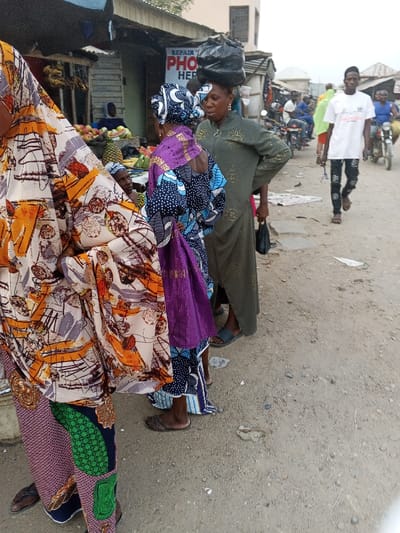 An outdoor scene in Zango-ndaji, Nigeria, depicts a busy street or market area under diffused daylight. The ground is an unpaved dirt road. In the immediate foreground on the left, a person is partially visible, wearing a patterned garment featuring orange, brown, grey, and black abstract designs over green patterned trousers. Immediately behind this person, a woman stands facing right, dressed in a blue and white patterned dress and a purple headwrap, holding a purple bag. To her right, a second woman stands, wearing a long olive-green dress with subtle golden patterns and balancing a large black plastic bag on her head while holding a smaller black bag in her left hand. These two women appear to be interacting. Behind them, a fruit stall displays items such as mangoes, pineapples, and watermelons, with a third person partially visible behind the stall. Above the stall, a sign reading "REPAIR PHONE HERE" is visible. Further into the background on the right, a young man walks away from the camera, wearing a white t-shirt and dark patterned trousers, with several other individuals and motorcycles visible further down the street, indicating ongoing traffic and activity. Buildings with corrugated metal roofing line the street, characteristic of market stalls or simple structures.