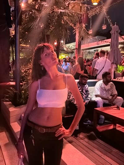 The image depicts an outdoor bar or restaurant scene at night in Pecatu, Indonesia, characterized by warm red and orange lighting. In the foreground, a light-skinned woman with dark hair and bangs stands slightly angled to the right, looking towards the viewer with her head tilted back and chin raised. She is wearing a white ribbed crop top with spaghetti straps, exposing her midriff, and dark pants with a brown belt. A set of keys hangs from her belt on her left hip. Her left arm is by her side, holding a white object, and her right hand rests on her right hip. She has earrings in both ears.
Behind her, the environment features numerous large palm trees illuminated by overhead lights. Several tables and seating areas are visible, occupied by numerous individuals. In the mid-ground, two men are seated on a light-colored sofa. The man on the left wears a black and white zebra-print shirt, while the man on the right wears a white, short-sleeved top with sparkling embellishments. A dark wooden table in front of them has sunglasses resting on it. Further in the background, a crowd of people is visible, some standing and some seated, with various lighting fixtures hanging above. The ground consists of wooden decking and concrete steps. An unbrella is visible on the right.