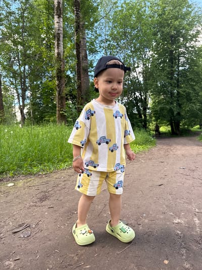 A young child stands on a dirt path in an outdoor setting in Rusanovo, Russia. The child, likely a boy, wears a black baseball cap, a yellow and white striped short-sleeved shirt, and matching shorts, both patterned with blue cartoon trucks that have "24" written on them. On his feet are bright lime green clogs, one of which features a small patch with "GRIN OUT" visible text, along with small charms including a white character and an orange basketball. He also wears a silver-colored bracelet on his left wrist.
The child looks directly towards the camera with a neutral to slightly smiling expression. The path extends into the background, flanked by lush green grass and numerous tall trees with dense green foliage, suggesting a park or forest environment. The sky is visible between the tree branches, indicating clear, bright daytime weather. The ground is a dry, textured dirt path with some scattered debris like twigs.