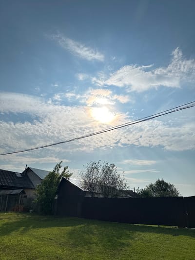 The image depicts a residential area in the village of Sredneye Kechevo, Russia, under a bright, partially clouded sky. The sun is visible, partially obscured by scattered white, puffy clouds that create a textured pattern across the blue sky. A halo-like effect is visible around the sun, suggesting atmospheric conditions. Two dark power lines stretch horizontally across the upper half of the frame. In the foreground, a well-maintained green lawn is visible, casting long shadows from unseen objects. Behind the lawn, a dark brown, solid fence runs across the width of the image. Beyond the fence, several residential buildings with visible roofs are present. One building on the left has a dark, corrugated metal roof and appears to be a house. Another building, further to the right, has a lighter-colored wall and a gray roof. Trees with green foliage are scattered between and behind the buildings. The overall impression is a rural or suburban landscape on a sunny day.