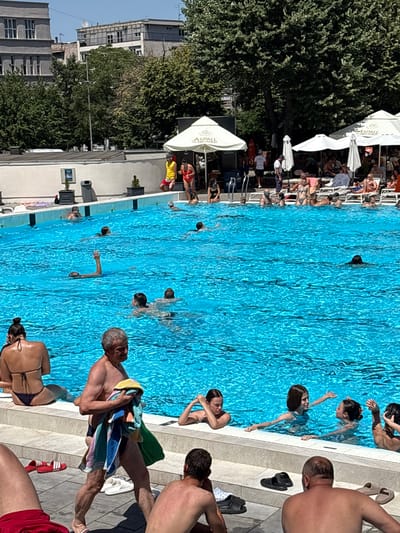 A group of people are enjoying a sunny day at an outdoor swimming pool, likely in Belgrade, Serbia. The pool water is a vibrant blue, reflecting the clear sky, suggesting it's a warm, bright day. Several individuals are swimming, while others are relaxing on lounge chairs or sitting by the poolside.

In the foreground, a man in swim trunks holds a colorful towel. Nearby, another man sits on the edge of the pool, facing away from the camera. A woman in a bikini is visible in the lower right of the frame. In the pool, a young girl with dark hair is reaching out a hand, and a younger child is swimming. Further out in the pool, several other people are visible, some submerged and others with their heads above water.

On the left side of the image, large green trees provide shade. Several white umbrellas and lounge chairs are scattered around the pool area, indicating a resort or public swimming facility. In the background, buildings are visible, one of which appears to be a multi-story apartment building.

There is some text visible on the white umbrellas: "ASSPALL" and "HIPS&WILD". The overall atmosphere appears relaxed and leisurely, consistent with a day at the pool during warm weather.