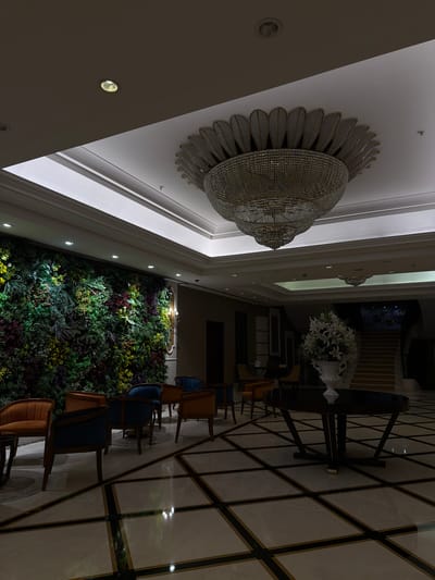 The image depicts the interior of a building, likely in Bağcılar, Turkey, featuring a blend of decorative elements and furniture. The perspective is angled upwards, capturing a large, ornate chandelier affixed to the ceiling. The ceiling itself is white, with recessed lighting visible.
To the right, a seating area is visible with chairs arranged around tables. The flooring has a geometric pattern created by dark lines against a light background. On the other side, there is a wall covered in greenery, resembling an indoor garden. In the background, near the center, a floral arrangement in a vase can be seen. No people are visible in the scene.