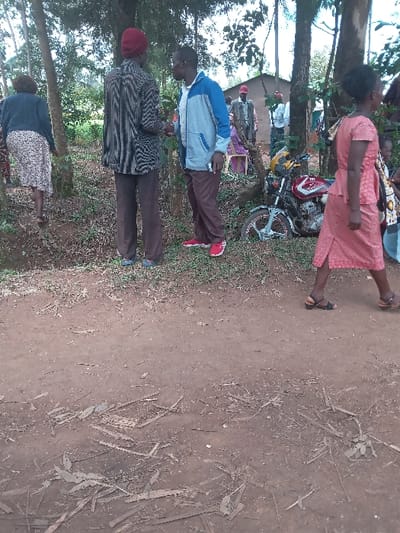 The image shows a group of people in a rural area located in Malava, Kenya at coordinates 0.498, 34.821. In the foreground, a dirt road with scattered leaves and twigs is seen. Two men are standing in the center of the image, one wearing a blue jacket and the other wearing a striped shirt and a red cap. The man in the blue jacket is holding a small object in his right hand and is talking to the man in the striped shirt. They both appear to be middle-aged.  Behind them, on the right, a woman wearing a pink dress and black sandals is walking away from the camera. She appears to be in her early 30s. A small, yellow motorcycle is parked on the left side of the image, behind the men. Behind the motorcycle, another woman is sitting on a chair, and several other people can be seen in the background. The environment is characterized by a dirt road, lush green vegetation, and a low-lying building behind the motorcycle.